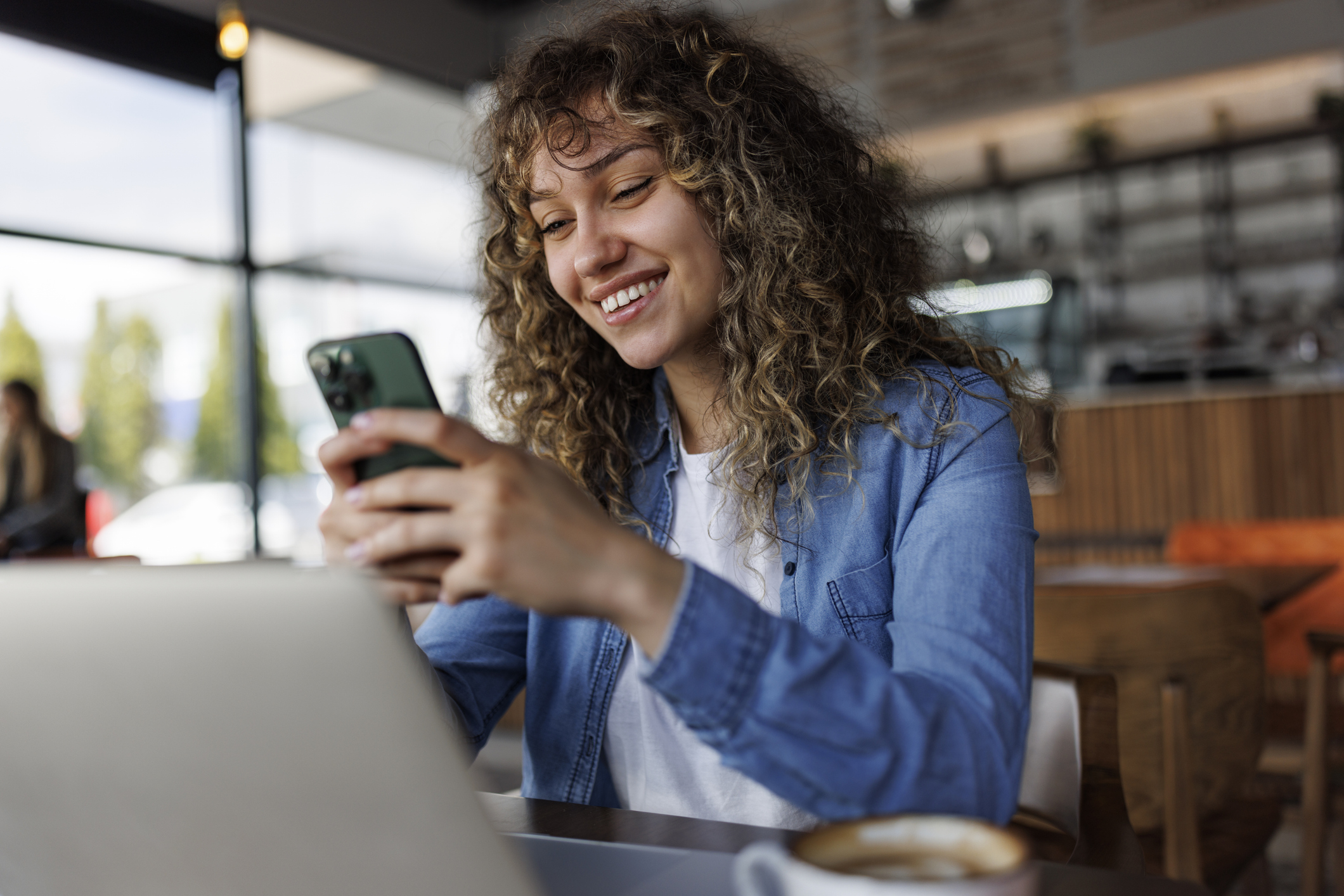 Woman smiling at phone in cafe while using laptop